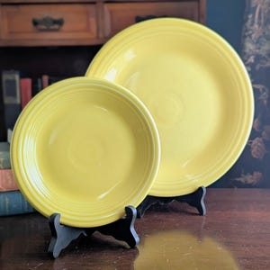 May include: Two bright yellow ceramic plates of different sizes, each featuring a series of concentric circular ridges along the rim. The plates are displayed upright on black plastic stands, showcasing a smooth, glossy finish and uniform color.