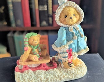 May include: A figurine featuring two golden-brown teddy bears on a snowy base. One bear wears a blue hooded coat with white trim and holds a small bell. The second bear wears a green striped hat and scarf while sitting on a red wooden sled.
