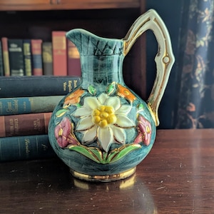 May include: A ceramic pitcher featuring a teal green base with a textured finish and a gold-toned handle and rim. The front displays a raised, three-dimensional white flower with a yellow center, accented by smaller orange and pink floral motifs.