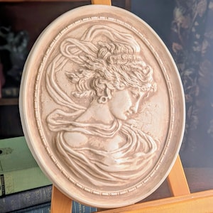 May include: Oval-shaped, off-white relief sculpture featuring a classical female profile with flowing hair and draped clothing. The sculpture is framed by a decorative border and mounted on a wooden easel. The artwork evokes a sense of classical elegance and historical art.