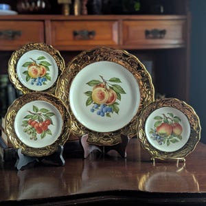 May include: A collection of vintage decorative plates with gold rims. Each plate features a different fruit design, including peaches, cherries, and apples, with green leaves and blue berries. The plates are displayed on wooden stands.