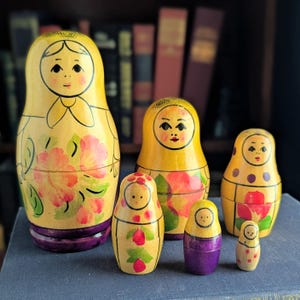 May include: A set of five yellow nesting dolls with hand-painted floral designs in pink and green. Each doll features a painted face with black eyes and a purple base. The dolls vary in size, with the largest on the left and the smallest on the far right.