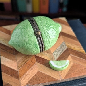 May include: A ceramic lime-shaped trinket box with a hinged, ornate metal band. The box is a vibrant green with a textured surface, and a small lime wedge is next to it. The box sits on a wooden surface with a chevron pattern.