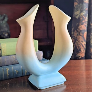 Könnte beinhalten: Eine Keramikvase in einer einzigartigen U-Form mit Farbverlauf. Die Vase wechselt von hellem Gelb oben zu einem sanften Blau am Boden. Sie hat einen quadratischen Sockel und ist wahrscheinlich ein dekorativer Gegenstand.