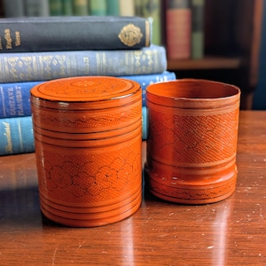 May include: Two orange lacquered wooden containers. One is a lidded cylinder with floral and geometric patterns. The other is a matching open cylinder. Both have black accent lines. Books are in the background.
