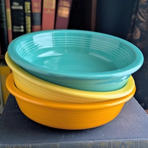 May include: A stack of three ceramic bowls in graduated sizes and vibrant colors. The top bowl is turquoise, the middle bowl is light yellow, and the bottom bowl is orange. Each bowl features a smooth, glossy finish and concentric ring detailing.