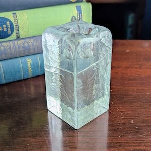 Puede incluir: Un bloque de vidrio transparente y texturizado con forma cuadrada y una parte superior ligeramente inclinada. El vidrio tiene un aspecto esmerilado y se encuentra sobre una superficie de madera oscura. Libros con cubiertas verdes, azules y doradas en el fondo.