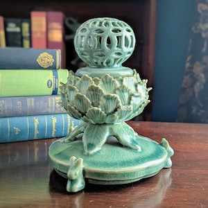 May include: A celadon green ceramic decorative object. The object features a spherical top with cut-out designs, a base shaped like an artichoke, and a circular base with small rabbit figurines. The piece is displayed on a wooden surface.