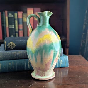 May include: A vintage ceramic pitcher with a handle. The pitcher features a green, yellow, and pink color scheme. The pitcher is sitting on a wooden surface with a stack of books in the background.
