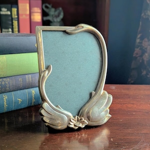 May include: A vintage brass picture frame with a swan design. The frame has a curved shape with swan heads at the top and swan wings at the bottom, with a small flower in the center. The frame holds a teal-colored backing and is displayed on a wooden surface.