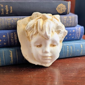 May include: A cream-colored, sculpted face of a child with closed eyes and a bow in the hair. The sculpture is set against a backdrop of antique books with blue and gold covers, creating a vintage aesthetic.