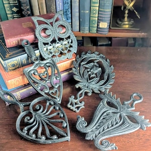 Puede incluir: Colección de salvamanteles decorativos de hierro fundido en varias formas, incluyendo un búho, un escudo con corazones y un diseño floral. Los salvamanteles son de color gris oscuro y están sobre una superficie de madera, con libros antiguos al fondo.