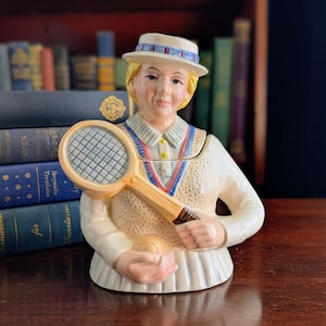 Könnte beinhalten: Eine Keramikfigur einer Tennisspielerin, die einen Schläger und einen Tennisball hält. Die Figur trägt ein weißes Tennisoutfit mit blau-roten Verzierungen und einen weißen Hut mit blau-rotem Band. Die Figur steht auf einer Holzoberfläche.