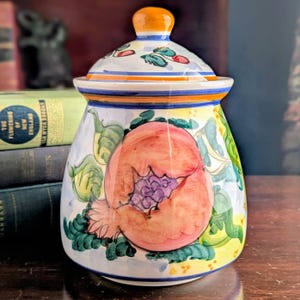 May include: A hand-painted ceramic jar with a lid. The jar features a design of a large, pink fruit with a cluster of purple grapes, surrounded by green leaves and vines. The lid and rim have blue and orange accents.