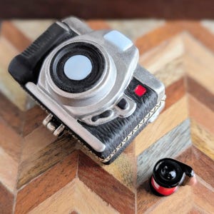 May include: A small, detailed camera figurine featuring a silver body with black textured accents, a prominent central lens with a light blue center, a small red square detail, and a separate, detached black and red cylindrical component nearby.
