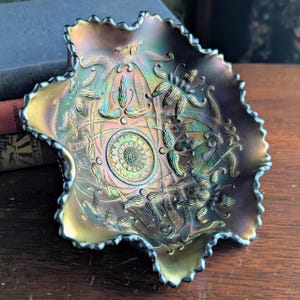 May include: A dark purple iridescent glass bowl with a ruffled, scalloped edge. The interior features a central circular medallion with a stippled texture, surrounded by embossed floral patterns and curved lines that shimmer with green and gold tones.