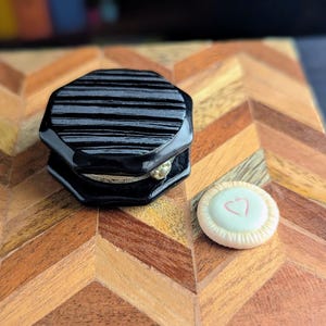 May include: A black octagonal container with a glossy, horizontally striped finish and a small silver-toned metal hinge, positioned next to a small, round, off-white button featuring a delicate pink heart outline on its center surface.
