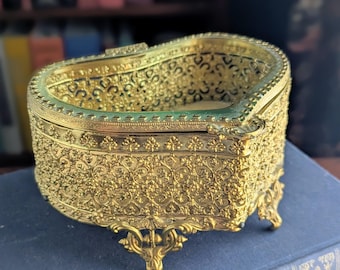 May include: A heart-shaped gold-tone metal trinket box featuring an intricate filigree pattern across the exterior walls. The container stands on four ornate, curved feet and includes a textured rim along the top edge with a light-colored interior.