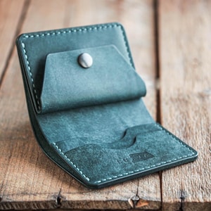 May include: Open teal leather wallet with a snap closure. The wallet has white stitching around the edges and features interior card slots. The wallet is displayed on a wooden surface.