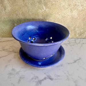 May include: A deep, vibrant blue ceramic berry bowl with a matching saucer. The bowl features a wide, flared rim and multiple circular drainage holes distributed across the bottom and lower sides, finished with a smooth, glossy glaze.
