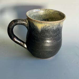 May include: A handmade ceramic mug with a black exterior that fades to a yellow-green rim. The mug has a rounded body and a textured handle. The interior is a light yellow-green color. This mug is perfect for coffee, tea, or other beverages.
