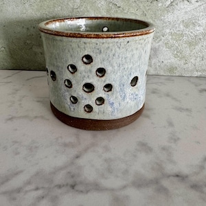 May include: A handmade ceramic container with a light blue and brown glaze. The cylindrical vessel features a series of circular holes and a brown rim and base. The pottery has a rustic, textured appearance, suitable for home decor.