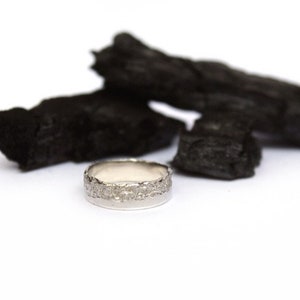 Puede incluir: Un anillo de plata con una banda texturizada. El anillo está sobre una pila de carbón vegetal negro.