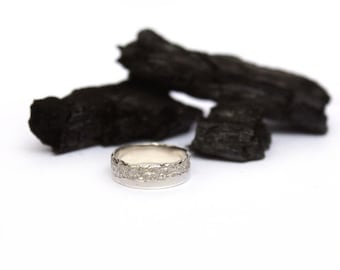 Könnte beinhalten: Ein silberner Ring mit einem strukturierten Band. Der Ring liegt auf einem Haufen schwarzer Holzkohle.