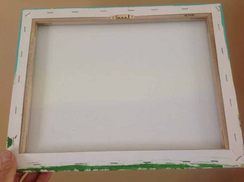 May include: The back of a stretched canvas painting with a wooden frame. The canvas is white and the frame is painted turquoise with green accents. The frame has a metal hanging hook.