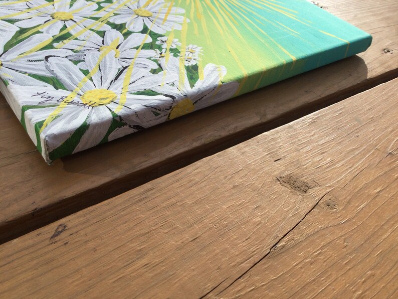 May include: A close-up of a canvas painting with a white daisy design. The background is a gradient of yellow, green, and blue. The painting is signed "Joy Russell" in the bottom left corner.