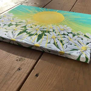 May include: A painting of a bright yellow sun with rays shining down on a field of white daisies. The background is a gradient of green and blue.