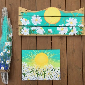 May include: Three pieces of art featuring a sun and daisies painted on wood and canvas. The top piece is a wooden sign with a turquoise background, a yellow sun, and white daisies. The middle piece is a canvas with a turquoise background, a yellow sun, and white daisies. The bottom piece is a piece of driftwood with a turquoise background, a yellow sun, and white daisies.