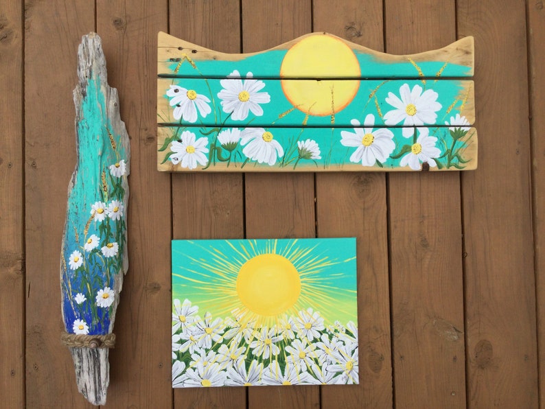 May include: Three pieces of art featuring a sun and daisies painted on wood and canvas. The top piece is a wooden sign with a turquoise background, a yellow sun, and white daisies. The middle piece is a canvas with a turquoise background, a yellow sun, and white daisies. The bottom piece is a piece of driftwood with a turquoise background, a yellow sun, and white daisies.