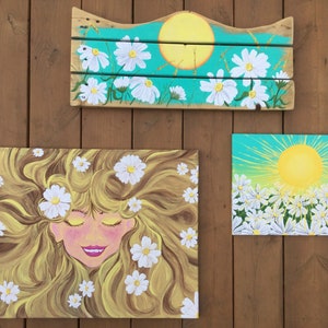 May include: Three paintings with a daisy theme. The first painting is a wooden sign with a yellow sun and white daisies on a turquoise background. The second painting is a canvas with a woman with long blonde hair and white daisies. The third painting is a canvas with a yellow sun and white daisies on a turquoise background.