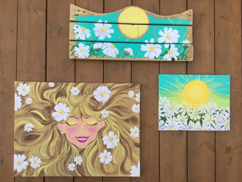 May include: Three paintings with a daisy theme. The first painting is a wooden sign with a yellow sun and white daisies on a turquoise background. The second painting is a canvas with a woman with long blonde hair and white daisies. The third painting is a canvas with a yellow sun and white daisies on a turquoise background.
