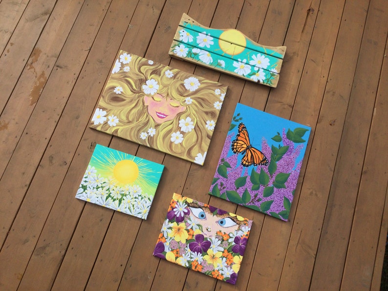 May include: Five paintings on canvas with floral and nature themes. One painting features a woman with blonde hair and white flowers, another has a butterfly on a purple flower, and another has a sun with white flowers. The remaining two paintings feature a woman's face with flowers.