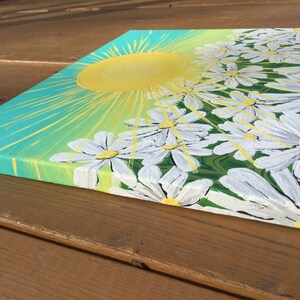 May include: A painting of a bright yellow sun with rays of light shining down on a field of white daisies with yellow centers. The background is a gradient of teal and green.