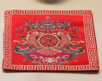 May include: A red and gold embroidered cushion with a decorative design. The cushion is placed on a white surface with a small white ceramic incense burner with a stick of incense.