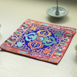 Peut inclure: Un coussin carré rouge, bleu et or avec des motifs complexes. Le coussin est sur une surface blanche avec un petit bol blanc et un brûle-encens en céramique.