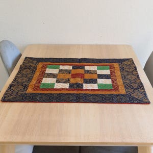 May include: A square table runner with a checkered pattern in shades of green, white, red, and gold. The runner has a dark blue border with gold accents and a red outer border.