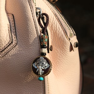 May include: A pink leather handbag with a zipper closure and a silver charm attached to the zipper pull. The charm features a detailed elephant design with turquoise accents.