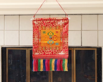 Könnte beinhalten: Ein roter Wandteppich mit goldenen und gelben Stickereien hängt an einer weißen Stange. Der Wandteppich zeigt ein farbenfrohes Mandala-Design mit dem Text "吉祥" in chinesischen Schriftzeichen. Der Wandteppich hat einen Fransenrand mit bunten Quasten.
