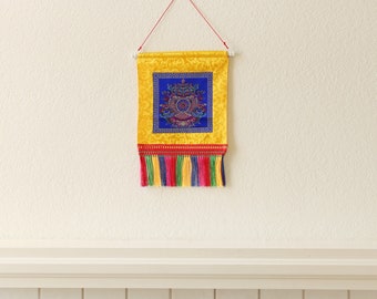 Könnte beinhalten: Ein gelber Wandteppich mit einem blauen und roten Mandala-Design hängt an einer weißen Wand über einem weißen gekachelten Kamin. Der Wandteppich hat einen Fransenbesatz aus bunten Quasten.