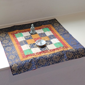 Può includere: Un tappetino da meditazione quadrato e fantasia con un design blu, oro, rosso e verde. Una piccola statua di Buddha grigia e un portaincenso bianco con un bastoncino di incenso sono sul tappetino. Il tappetino è su una superficie bianca.