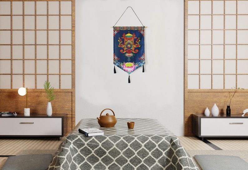 May include: A blue and gold tapestry with a floral design and tassels hanging from the bottom. The tapestry is hanging on a white wall in a room with a wooden floor and a table with a teapot and a cup on it.