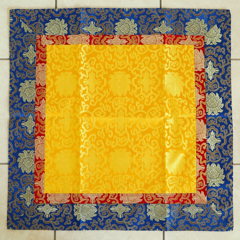 Puede incluir: Un coj&iacute;n cuadrado amarillo con un borde azul y rojo. El coj&iacute;n tiene un patr&oacute;n floral y est&aacute; hecho de una tela suave y lujosa.