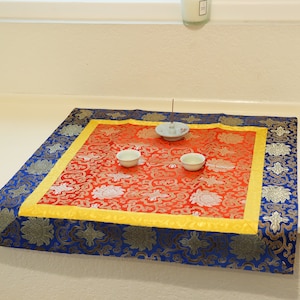 Puede incluir: Un coj&iacute;n cuadrado rojo y dorado con un borde azul. Dos cuencos peque&ntilde;os blancos y un porta incienso blanco se colocan en el coj&iacute;n.