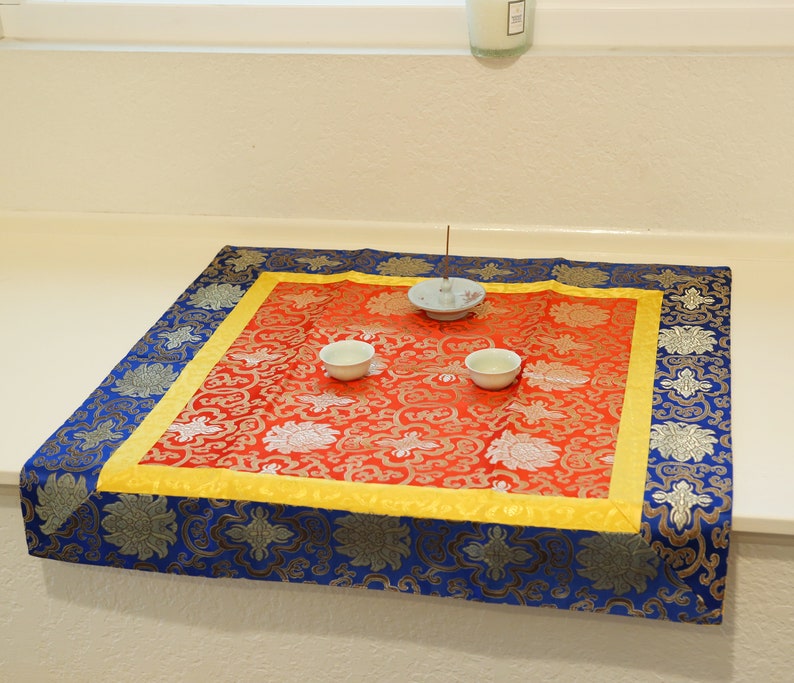 Puede incluir: Un coj&iacute;n cuadrado rojo y dorado con un borde azul. Dos cuencos peque&ntilde;os blancos y un porta incienso blanco se colocan en el coj&iacute;n.