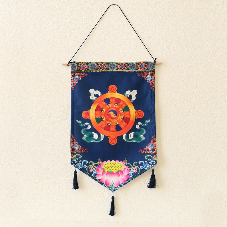 May include: A blue tapestry with a colorful wheel of dharma design, surrounded by clouds and flowers. The tapestry has tassels on the bottom and is hanging from a wooden dowel.