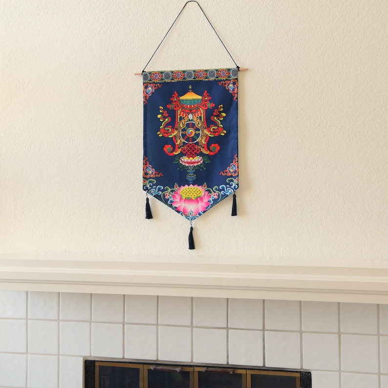 May include: A blue tapestry with a colorful mandala design featuring a wheel of dharma, lotus flowers, and intricate patterns. The tapestry is hanging on a white wall with a white mantelpiece below.
